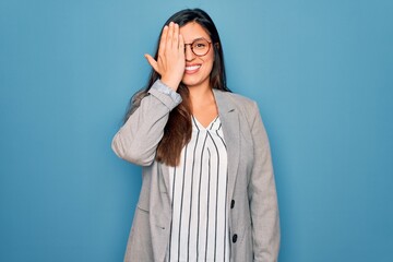 Sticker - Young hispanic business woman wearing glasses standing over blue isolated background covering one eye with hand, confident smile on face and surprise emotion.