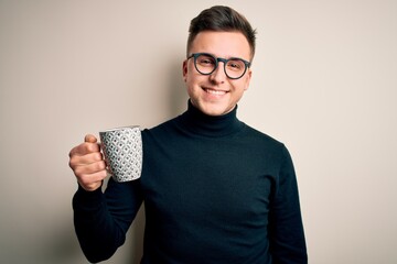 Young handsome caucasian man wearing glasses drinking a cup of hot coffee with a happy face standing and smiling with a confident smile showing teeth