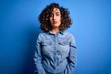 Poster - Young beautiful curly arab woman wearing casual denim shirt standing over blue background Relaxed with serious expression on face. Simple and natural looking at the camera.