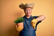 Middle age hoary farmer man wearing apron and hat holding plant pot over yellow background with angry face, negative sign showing dislike with thumbs down, rejection concept
