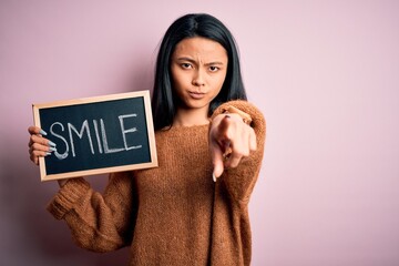 Wall Mural - Young chinese woman holding blackboard with smile word over isolated pink background pointing with finger to the camera and to you, hand sign, positive and confident gesture from the front