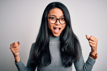 Wall Mural - Young beautiful chinese woman wearing glasses and sweater over isolated white background celebrating surprised and amazed for success with arms raised and open eyes. Winner concept.