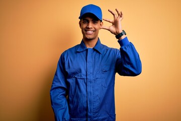 Wall Mural - Young african american mechanic man wearing blue uniform and cap over yellow background smiling and confident gesturing with hand doing small size sign with fingers looking and the camera. Measure