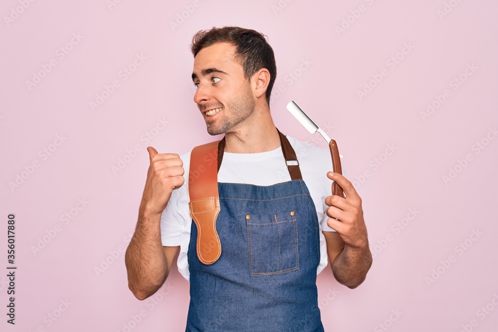 Handsome barber man with blue eyes wearing apron using razor blade over pink background pointing and showing with thumb up to the side with happy face smiling - obrazy, fototapety, plakaty 