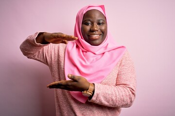 Poster - Young african american plus size woman wearing muslim hijab over isolated pink background gesturing with hands showing big and large size sign, measure symbol. Smiling looking at the camera. Measure