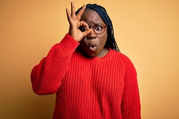 Sticker - African american plus size woman with braids wearing casual sweater over yellow background doing ok gesture shocked with surprised face, eye looking through fingers. Unbelieving expression.