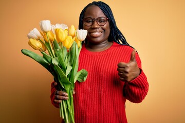 Poster - Young african american plus size woman with braids holding bouquet of yellow tulips flower doing happy thumbs up gesture with hand. Approving expression looking at the camera showing success.