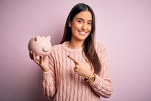 Young Beautiful Brunette Woman Holding Piggy Bank Saving Money For Retirement Very Happy Pointing With Hand And Finger