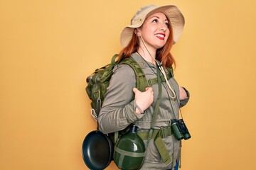 Poster - Young redhead backpacker woman hiking wearing backpack and hat over yellow background looking away to side with smile on face, natural expression. Laughing confident.