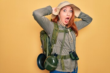 Wall Mural - Young redhead backpacker woman hiking wearing backpack and hat over yellow background Crazy and scared with hands on head, afraid and surprised of shock with open mouth