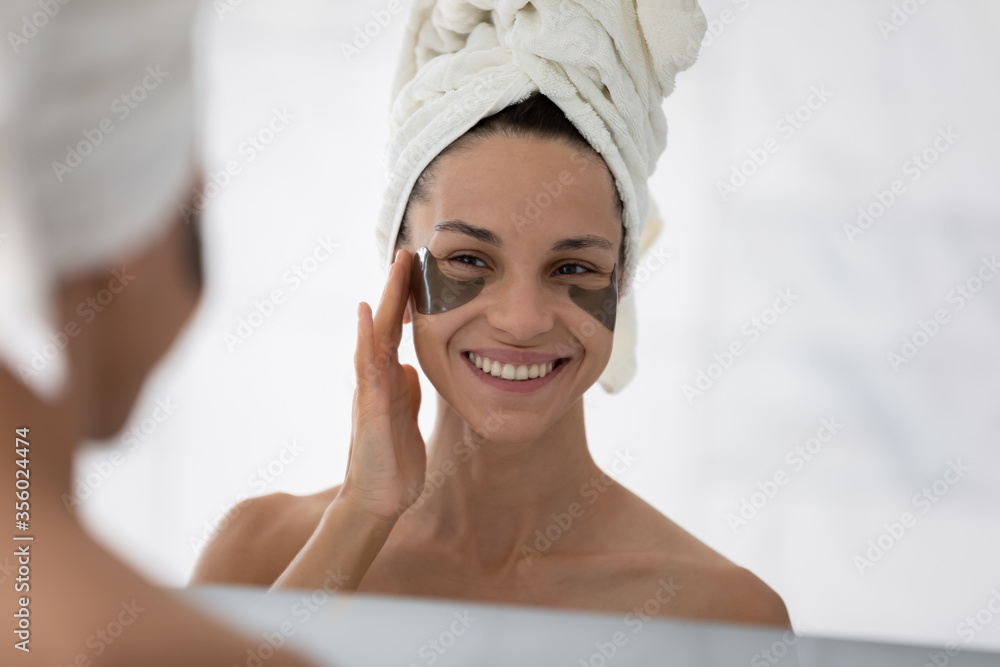 Mirror reflection smiling woman wearing white bath towel on head applying hydrogel eye care patches, standing in bathroom, moisturizing skin under eyes, enjoying skincare procedures - obrazy, fototapety, plakaty 