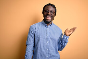 Wall Mural - Young handsome african american man wearing shirt and glasses over yellow background smiling cheerful presenting and pointing with palm of hand looking at the camera.