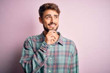 Poster - Young handsome man with beard wearing casual shirt standing over pink background with hand on chin thinking about question, pensive expression. Smiling with thoughtful face. Doubt concept.