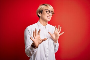 Poster - Young blonde business woman with short hair wearing glasses over red background disgusted expression, displeased and fearful doing disgust face because aversion reaction. With hands raised