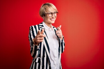 Wall Mural - Blonde business woman with short hair wearing glasses and striped jacket over red background pointing fingers to camera with happy and funny face. Good energy and vibes.