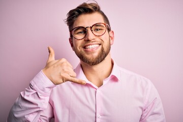 Wall Mural - Young handsome blond man with beard and blue eyes wearing pink shirt and glasses smiling doing phone gesture with hand and fingers like talking on the telephone. Communicating concepts.