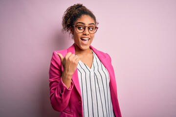 Poster - Beautiful african american businesswoman wearing jacket and glasses over pink background smiling with happy face looking and pointing to the side with thumb up.