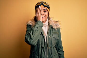 Sticker - Young african american afro skier girl wearing snow sportswear and ski goggles covering one eye with hand, confident smile on face and surprise emotion.