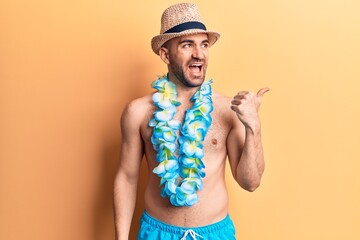 Young handsome bald man shirtless wearing swimwear and hawaiian lei pointing thumb up to the side smiling happy with open mouth