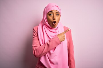 Wall Mural - Young African American afro woman wearing muslim hijab over isolated pink background Surprised pointing with finger to the side, open mouth amazed expression.