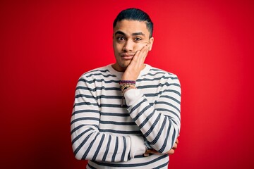 Wall Mural - Young brazilian man wearing casual striped t-shirt standing over isolated red background thinking looking tired and bored with depression problems with crossed arms.