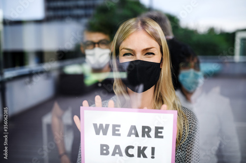 Woman with face mask back at work in office after lockdown, holding we are back sign.
