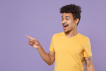 Wall Mural - Amazed young african american guy in casual yellow t-shirt posing isolated on pastel violet wall background studio portrait. People lifestyle concept. Mock up copy space. Pointing index finger aside.