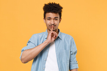 Wall Mural - Secret young african american guy in casual blue shirt posing isolated on yellow background studio. People lifestyle concept. Mock up copy space. Saying hush be quiet with finger on lips shhh gesture.