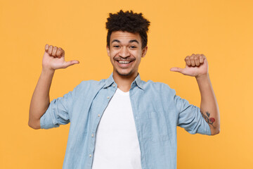 Wall Mural - Cheerful young african american guy in casual blue shirt posing isolated on yellow background studio portrait. People sincere emotions lifestyle concept. Mock up copy space. Point thumbs on himself.