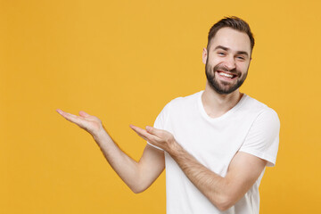 Smiling young bearded man guy in white casual t-shirt posing isolated on yellow wall background studio portrait. People sincere emotions lifestyle concept. Mock up copy space. Pointing hands aside.