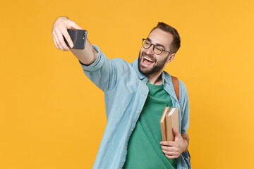 Wall Mural - Funny young man student in casual clothes glasses with backpack hold books isolated on yellow background studio. Education in high school university college concept. Doing selfie shot on mobile phone.