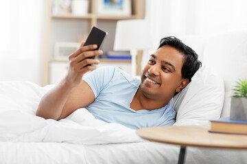 Wall Mural - people, bedtime and rest concept - happy smiling indian man with smartphone lying in bed at home