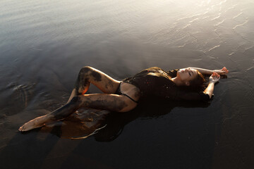 Wall Mural - Sexy woman wearing black tunic is posing on the beach with black sand