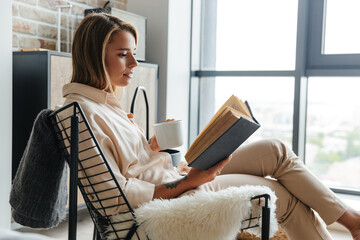 Poster - Image of woman drinking tea and reading book while sitting on armchair