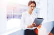 © BullRun - Young serious businesswoman using touch pad while standing in office interior near window