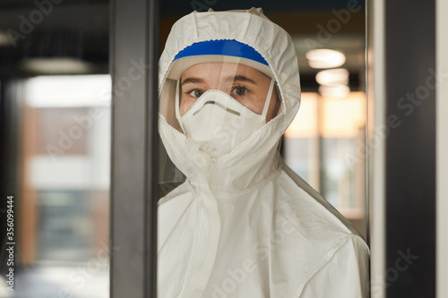 Waist up portrait of female worker wearing protective suit looking at camera while cleaning glass windows indoors during disinfection, copy space