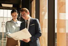 Waist Up Portrait Of Smiling Rental Agent Showing Floor Plans To Female Client While Standing In Office Building Interior Lit By Sunlight, Copy Space
