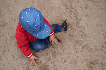 one year old boy wearing a cap, red jacket, blue pants and sneaker is sitting in the sand