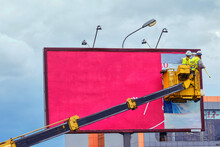 Two Workers Install Red Billboard On Roadside Of City Street