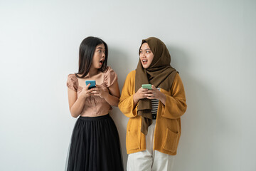 Wall Mural - two friendly girls look at each other and express shock at each other when using their smartphone with an isolated background