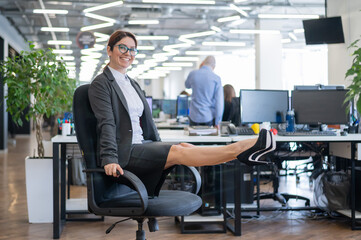 Happy business woman doing abdominal muscle exercises in an open space office. A red-haired smiling female employee in a skirt and high heels is dying off a chair in the workplace. Fitness at work.