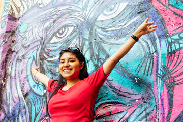 Young latin brunette girl with sunglasses in her hair posing with outstretched arms on wall with graffiti with eyes