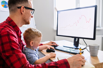 Father works from home office while babysitting with toddler kid. Dad sits at the desk with a little son on his laps and typing on keyboard