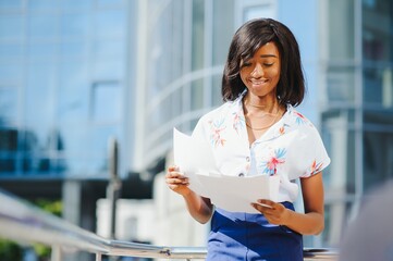 Wall Mural - Content formal African American woman in suit standing on sidewalk on street and reading papers in file against contemporary building