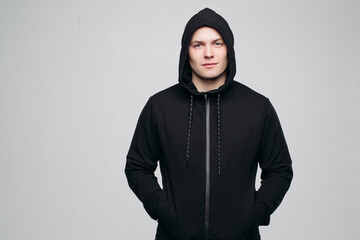 Front view of serious athletic man in black shirt with hood on head, posing, holding hands in pockets. Handsome and stylish male looking at camera. Concept of male fashion and look.