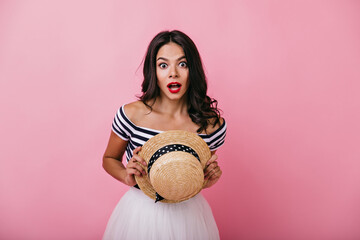 Wall Mural - Amazed brown-haired woman looking to camera with mouth open. Spectacular caucasian girl with straw hat in hands standing on pink background.