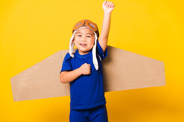 Wall Mural - Happy Asian handsome funny child or kid little boy smile wear pilot hat play and goggles raise hand up with toy cardboard airplane wings flying, studio shot isolated yellow background, Startup freedom