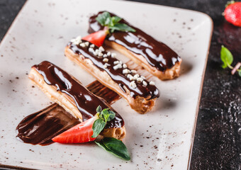 Wall Mural - Homemade cake eclairs or profiteroles with custard, chocolate and strawberries on dark background served with cup of coffee. Traditional french eclairs, dessert. Closeup