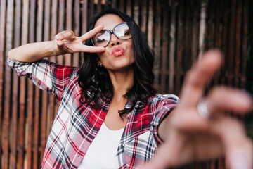 Wall Mural - Jocund woman in casual outfit playfully making selfie. Attractive tanned girl fooling around on wooden background.