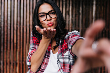 Wall Mural - Lovable european woman in glasses sending air kiss to camera. Elegant female model in checkered shirt making selfie in front on wooden fence.